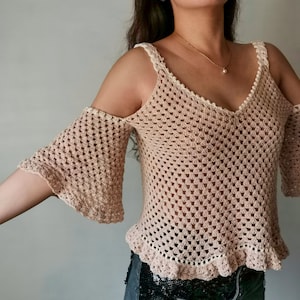 May include: A beige crochet top with a white trim. The top has a V-neck and is sleeveless with a cold shoulder design. The top has a ruffled hem.