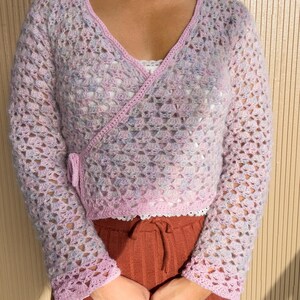 K&ouml;nnte beinhalten: Ein hellrosa, offen gewebter H&auml;kel-Wickelpullover mit langen &Auml;rmeln. Der Pullover hat einen V-Ausschnitt und ist mit einer dunkleren rosa Bord&uuml;re versehen. Die &Auml;rmel und der Saum sind mit einem gewellten Rand versehen. Der Pullover wird &uuml;ber einem wei&szlig;en Oberteil getragen und mit rostfarbenen Hosen kombiniert.