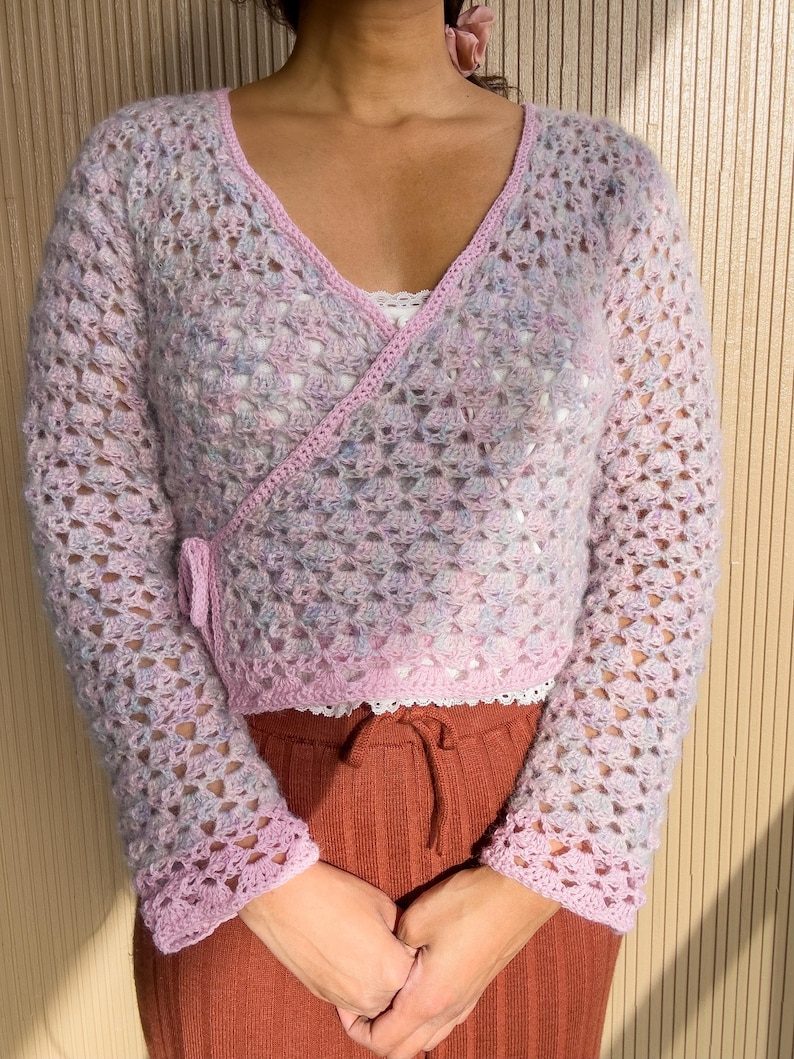 K&ouml;nnte beinhalten: Ein hellrosa, offen gewebter H&auml;kel-Wickelpullover mit langen &Auml;rmeln. Der Pullover hat einen V-Ausschnitt und ist mit einer dunkleren rosa Bord&uuml;re versehen. Die &Auml;rmel und der Saum sind mit einem gewellten Rand versehen. Der Pullover wird &uuml;ber einem wei&szlig;en Oberteil getragen und mit rostfarbenen Hosen kombiniert.