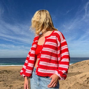 Op de afbeelding: Een rood en roze gestreept gehaakt vest met lange mouwen en knoopsluitingen. Het vest wordt gecombineerd met een rode top en lichtblauwe jeans. De achtergrond toont een blauwe lucht en een zandlandschap.