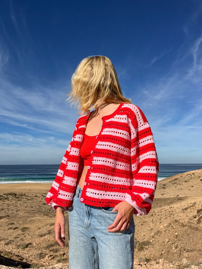 Op de afbeelding: Een rood en roze gestreept gehaakt vest met lange mouwen en knoopsluitingen. Het vest wordt gecombineerd met een rode top en lichtblauwe jeans. De achtergrond toont een blauwe lucht en een zandlandschap.