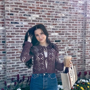 Pu&ograve; includere: Una donna indossa un cardigan di pizzo viola con un dettaglio a cravatta, jeans blu e una borsa tote beige con la scritta "Chlo&eacute;" stampata su di essa. Tiene un caff&egrave; freddo nella mano destra.