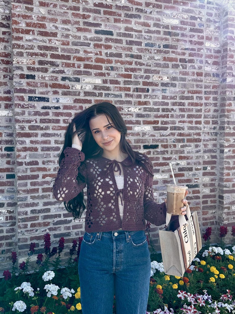 Pu&ograve; includere: Una donna indossa un cardigan di pizzo viola con un dettaglio a cravatta, jeans blu e una borsa tote beige con la scritta "Chlo&eacute;" stampata su di essa. Tiene un caff&egrave; freddo nella mano destra.