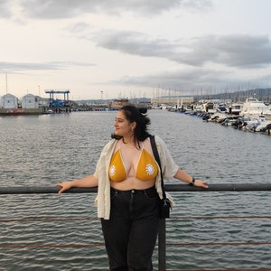 Puede incluir: Una mujer con un top de bikini de crochet amarillo con detalles de flores blancas, una camisa de lino blanca, pantalones vaqueros negros y zapatillas blancas. Est&aacute; de pie en un muelle con vistas a un puerto con barcos en el fondo.