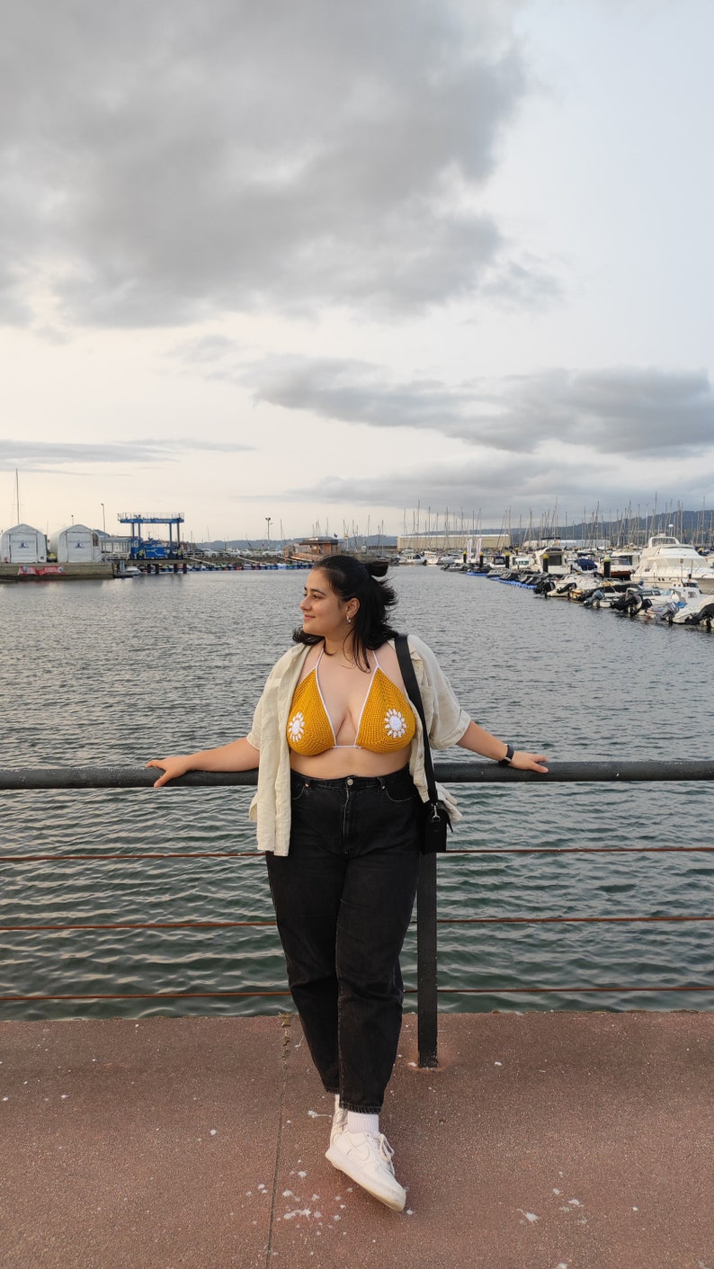 Puede incluir: Una mujer con un top de bikini de crochet amarillo con detalles de flores blancas, una camisa de lino blanca, pantalones vaqueros negros y zapatillas blancas. Est&aacute; de pie en un muelle con vistas a un puerto con barcos en el fondo.