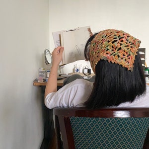 May include: A person wearing a crocheted bandana in shades of orange, green, and brown, sitting in a chair with a green and white patterned upholstery. The person is reading a book.