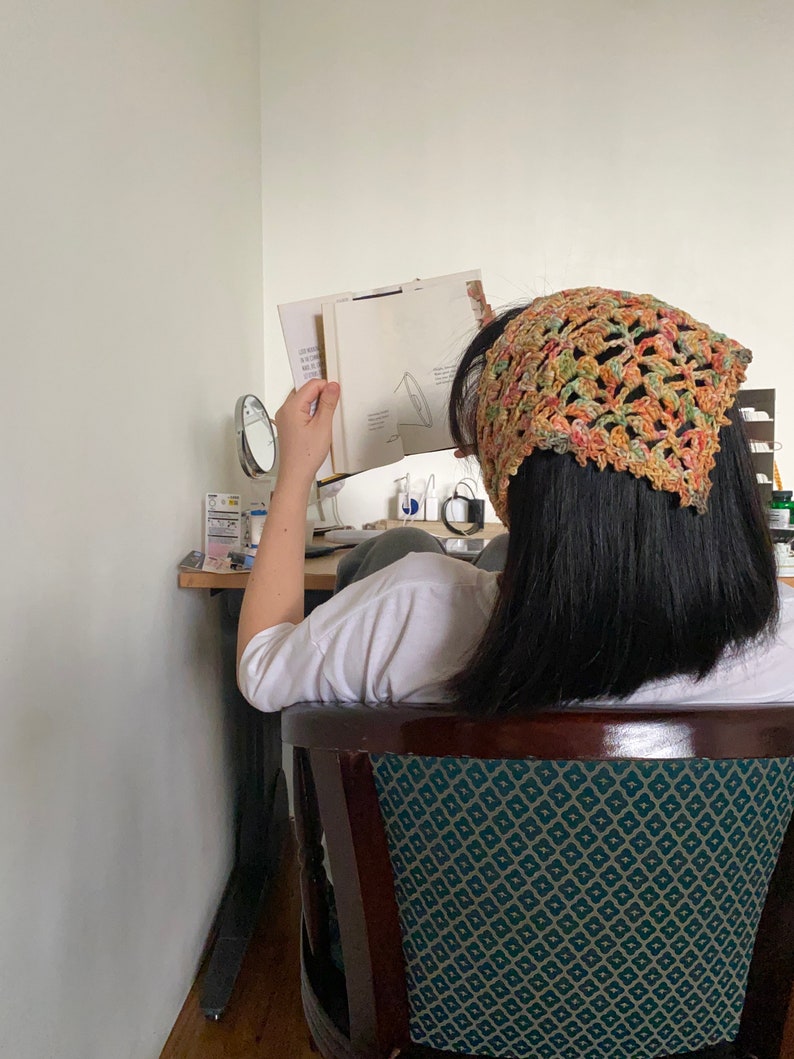 May include: A person wearing a crocheted bandana in shades of orange, green, and brown, sitting in a chair with a green and white patterned upholstery. The person is reading a book.