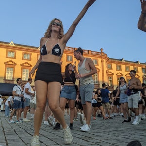Puede incluir: Una mujer con un top de bikini negro con adornos de flores blancas, una minifalda negra y zapatillas blancas baila con los brazos en alto. Est&aacute; rodeada de otras personas en una multitud.