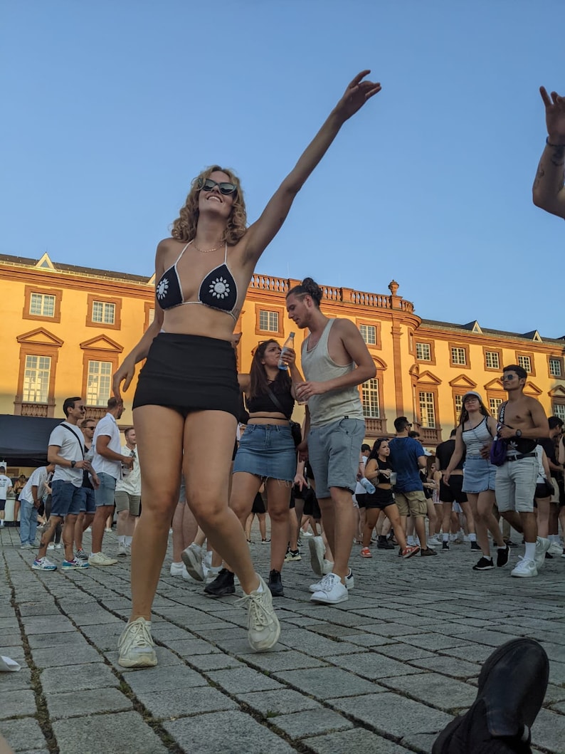 Puede incluir: Una mujer con un top de bikini negro con adornos de flores blancas, una minifalda negra y zapatillas blancas baila con los brazos en alto. Est&aacute; rodeada de otras personas en una multitud.