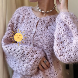 Könnte beinhalten: Ein helllilafarbener Häkelcardigan mit Knopfleiste und langen Ärmeln. Der Cardigan hat ein zartes, durchbrochenes Muster und eine gehäkelte Sonne mit dem Text "CROCHET PDF PATTERN" in der linken unteren Ecke.