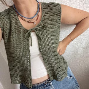 Op de afbeelding: Een groen gebreid vest zonder mouwen met een strik aan de voorkant. Het vest wordt gedragen over een wit tanktop en een blauwe jeans.