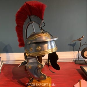 May include: A Roman-style helmet with a red plume, silver-coloured metal, and gold-coloured trim. The helmet is displayed on a wooden stand. The text "TYAGI EXPORT India" is visible on the stand.