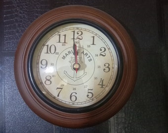May include: Round wall clock with a brown wooden frame and a cream-colored face. The clock face features black numerals, a nautical anchor design, and the words "MARINE ARTS". The clock hands point to 12:00.