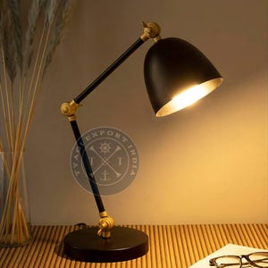 Puede incluir: Una lámpara de escritorio negra con un brazo ajustable dorado y una base redonda. La lámpara tiene una pantalla negra con interior dorado. La lámpara está encendida, emitiendo una luz cálida. La lámpara está sobre una superficie de madera.