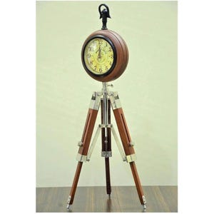 May include: A decorative clock on a tripod stand. The clock face is round, with a dark wood surround and a yellow face. The tripod legs are made of wood and metal. The clock's height is approximately 100 cm.
