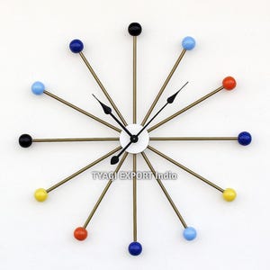 May include: A mid-century modern wall clock with a white face and black hands. The clock features brass-colored rods with colorful spherical accents in blue, red, yellow, and black. The clock's diameter is approximately 18 inches.