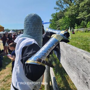 以下が含まれることがあります： 中世の鎧を着た人。鎖かたびらのフードと金属製の腕当てが含まれています。腕当ては銀色で、金色のアクセントと革のストラップが付いています。その人は、晴れた日に木の柵の近くに立っています。