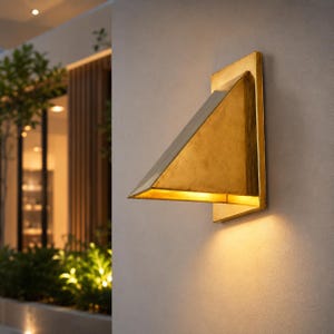 May include: A wall-mounted light fixture with a triangular, gold-toned metal shade. The fixture is attached to a rectangular backplate and emits a warm, downward-facing glow against the wall surface, creating a distinct geometric silhouette.