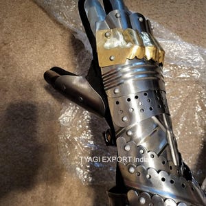 以下が含まれることがあります： 鋼と真鍮で作られた中世のガントレットのクローズアップ。ガントレットは、関節式の指と、リベットと装飾的なエッジを備えた詳細なデザインが特徴です。真鍮のアクセントが鎧にエレガントさを加えています。