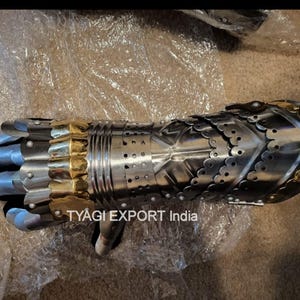 以下が含まれることがあります： 金色のアクセントが付いた銀色の金属製ガントレット。ガントレットは重なり合うプレートでできており、詳細なデザインが施されています。「TYAGI EXPORT India」の文字が見えます。