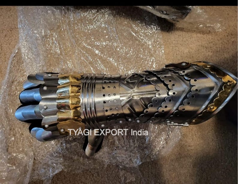 以下が含まれることがあります： 金色のアクセントが付いた銀色の金属製ガントレット。ガントレットは重なり合うプレートでできており、詳細なデザインが施されています。「TYAGI EXPORT India」の文字が見えます。