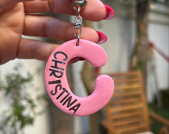 May include: A pink letter "C" keyring with the name "CHRISTINA" in black lettering. The keyring is attached to a silver clasp. The background is blurred, showing a green outdoor setting.