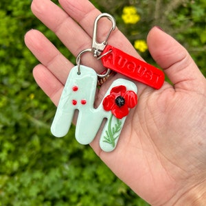 May include: A handmade keychain featuring a light green letter "M" with red dots and a red poppy flower. A red tag with the word "AUGUST" is attached. The keychain has a silver clasp.