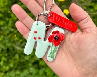 May include: A handmade keychain featuring a light green letter "M" with red dots and a red poppy flower. A red tag with the word "AUGUST" is attached. The keychain has a silver clasp.