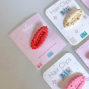 Puede incluir: Varias pinzas para el pelo con nombres se muestran en tarjetas rosas. Las pinzas son de varios colores, incluyendo rojo, rosa y crema, con nombres como "Maria", "Nour" y "Yasma". Las tarjetas tienen la marca "Bekle" y la frase "Hair Clips".