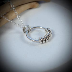 May include: A silver-coloured necklace featuring a delicate chain and a circular pendant. The pendant consists of a larger silver ring adorned with six smaller, smooth silver rings that slide along the circumference of the main circular frame.