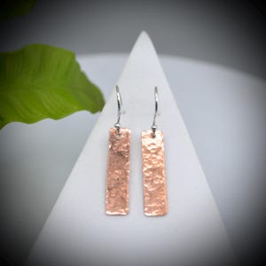 May include: Two copper-coloured rectangular earrings with a hammered texture. They hang from silver-coloured ear wires. The earrings are displayed against a white background with a green leaf in the upper left corner.