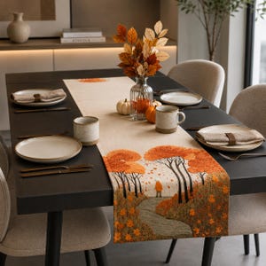 May include: A rectangular beige table runner featuring an embroidered autumn landscape with orange trees, a winding path, and a person in an orange coat. The runner is placed on a dark table with ceramic plates, mugs, and napkins.