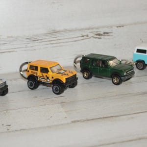 May include: Four miniature toy SUV keychains in various colors: silver, yellow, green, and light blue. Each keychain features a detailed replica of a classic SUV design, complete with wheels and a keyring attachment. The vehicles are arranged in a row.