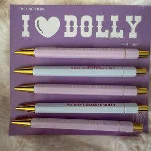 May include: A pen set with four pens in light pink and white with gold tips. The pens have text that reads "I Dolly", "What Would Dolly Do?", "Dolly for President", and "We Don't Deserve Dolly". The pen set is on a purple background.
