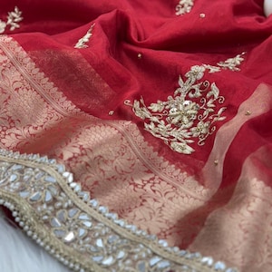 Puede incluir: Primer plano de una tela roja vibrante con intrincados bordados y adornos dorados. La tela presenta un detallado patrón floral a lo largo del borde y disperso por toda la superficie, con pequeños acentos espejados. El diseño sugiere un textil lujoso.