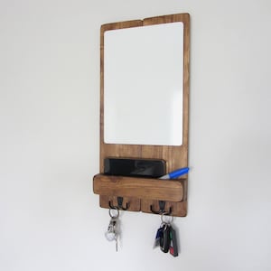 May include: A wooden wall organiser with a white dry-erase board, a shelf for a phone, and key hooks. A blue marker and keys hang from the hooks. The wood has a warm brown stain.