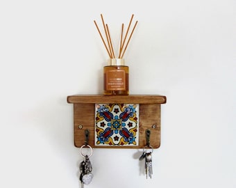 May include: A wooden key holder with a decorative tile insert featuring a colorful floral pattern. The key holder has two hooks and a small shelf holding a glass bottle of essential oil diffuser with reeds.