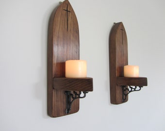 Könnte beinhalten: Zwei Wandleuchter aus Holz mit dunklem Finish, jeweils mit einem Kreuz-Schnitzmuster oben. Ein kleines Regal hält eine brennende, weiße Kerze. Die Leuchter sind mit dekorativen Metallhalterungen an einer weißen Wand befestigt.