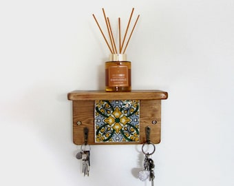 May include: A wooden key holder with a decorative tile insert featuring a floral pattern in yellow, blue, and green. The key holder has two hooks and a small shelf holding a glass bottle with reeds.