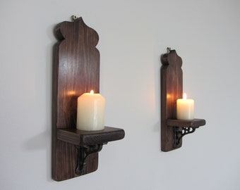 Könnte beinhalten: Zwei dunkelbraune Wandleuchter aus Holz mit dekorativer Oberseite und kleinem Regal. Jeder Leuchter enthält eine brennende weiße Kerze. Die Leuchter haben schwarze Metallhalterungen und sind an einer weißen Wand befestigt. Die Kerzen sind angezündet und werfen einen warmen Schein.