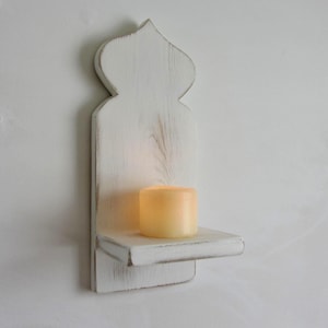 Op de afbeelding: Een witte houten wandkandelaar met een kaars. De kandelaar heeft een decoratieve bovenkant en een klein plankje om de kaars op te plaatsen. De kaars heeft een warme gele kleur en het hout heeft een verweerde afwerking.