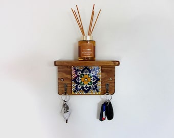 May include: A wooden key holder with a shelf, featuring a decorative tile with blue, yellow, and green floral patterns. It has two hooks with keys hanging from them and a brown glass diffuser bottle with reeds.