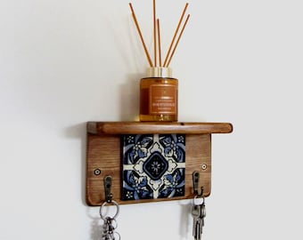 May include: A wooden key holder with a shelf, featuring a decorative blue and white tile. The key holder has two hooks with keys hanging from them. A glass bottle with reeds sits on the shelf.