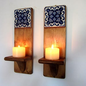 Puede incluir: Dos apliques de pared de madera con una vela encendida cada uno. Cada aplique tiene una baldosa decorativa azul y blanca en la parte superior. Las velas emiten un brillo cálido. Los apliques son de madera.
