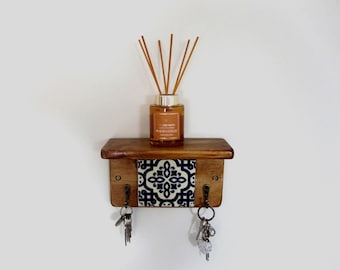 May include: A small, wooden shelf with a decorative blue and white tile insert. The shelf holds a brown glass diffuser bottle with reeds and a set of keys hanging below. The shelf has two hooks for keys.