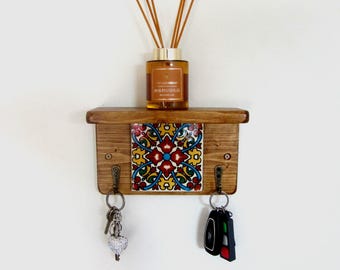 May include: A wooden key holder with a shelf, featuring a colorful patterned tile. A glass bottle with reeds sits on the shelf. Keys hang from metal hooks below. The key holder is mounted on a white wall.
