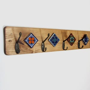 May include: A wooden coat rack with six black metal hooks. The rack features decorative square tiles with colourful floral patterns in blue, orange, and green. The wood has a natural finish.