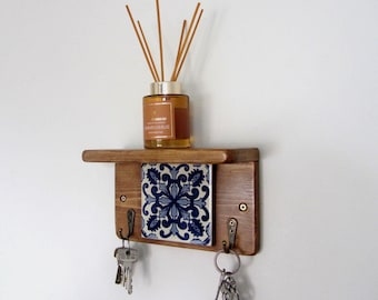 May include: A wooden key holder with a shelf, a decorative blue and white tile, and two hooks. A glass bottle with reeds sits on the shelf. Keys hang from the hooks. The key holder is mounted on a white wall.