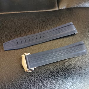 May include: Two-piece dark blue watch strap with a silver-colored clasp. The strap is made of a flexible material with horizontal grooves. One piece has several holes for adjustment.
