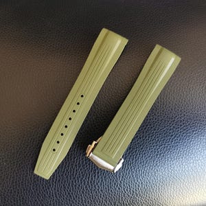 May include: Two olive green watch straps with a textured surface and vertical lines. One strap has several holes for adjustment. The other strap has a silver clasp. The straps are displayed on a dark, textured surface.